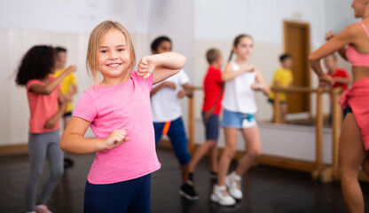 Fototapeta na wymiar Smiling little girl training movements of vigorous dance with group of tweens in children dance studio