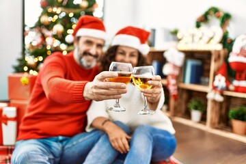 Middle age hispanic couple wearing christmas hat. Sitting on the floor toasting with champagne at home.