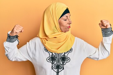 Middle age hispanic woman wearing traditional islamic hijab scarf showing arms muscles smiling...