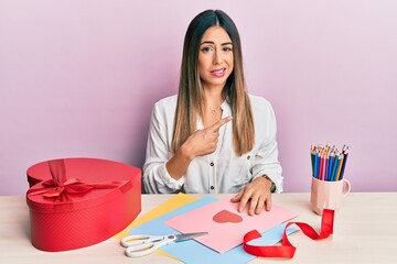 Young hispanic woman making valentine gift sitting on the table pointing aside worried and nervous...