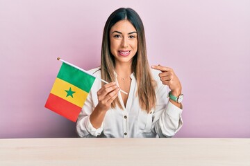 Young hispanic woman holding senegal flag sitting on the table smiling happy pointing with hand and...