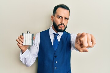 Young man with beard drinking whiskey from flask pointing with finger to the camera and to you, confident gesture looking serious