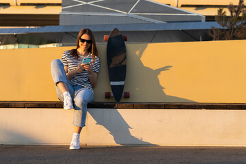 Millennial woman chat in mobile phone use smartphone application sitting outdoors with longboard skate in park. Smiling girl skater dressed in casual clothes texting or surfing web on street at sunset