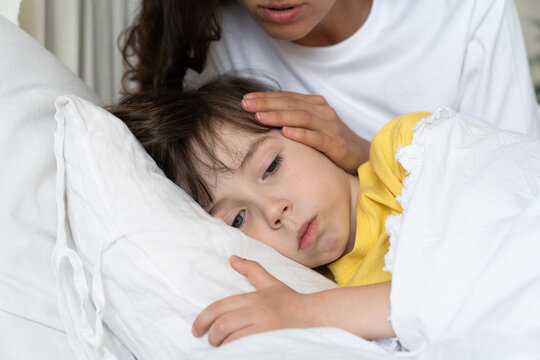 Closeup portrait of sick kid lying in bed with worried mom on bedside comfort child suffering from fever or flu. Female parent caring of exhausted toddler boy. Toddler sickness and home cure concept