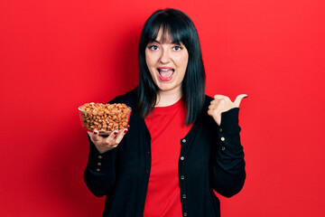 Young hispanic woman holding peanuts pointing thumb up to the side smiling happy with open mouth