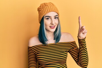 Young modern girl wearing wool hat showing and pointing up with finger number one while smiling confident and happy.