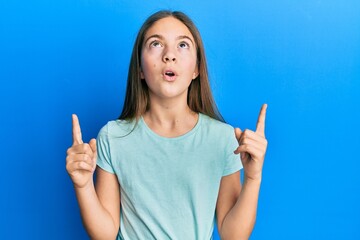 Beautiful brunette little girl wearing casual white t shirt amazed and surprised looking up and pointing with fingers and raised arms.