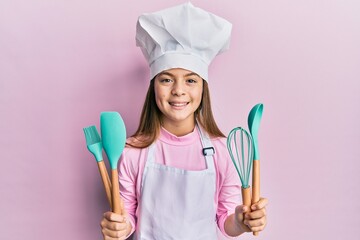 Beautiful brunette little girl wearing professional cook apron holding cooking tools smiling with a...