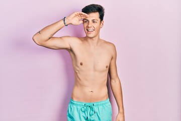 Fototapeta premium Young hispanic man wearing swimwear shirtless very happy and smiling looking far away with hand over head. searching concept.