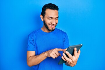 Hispanic man with beard using touchpad device looking positive and happy standing and smiling with a confident smile showing teeth