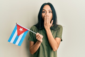 Young hispanic girl holding cuba flag covering mouth with hand, shocked and afraid for mistake....