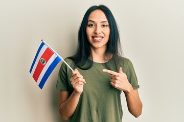 Young hispanic girl holding costa rica flag smiling happy pointing with hand and finger - obrazy, fototapety, plakaty