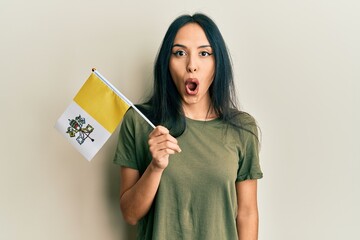 Young hispanic girl holding vatican city flag scared and amazed with open mouth for surprise,...