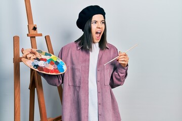 Young brunette woman standing by empty easel stand holding palette angry and mad screaming frustrated and furious, shouting with anger. rage and aggressive concept.