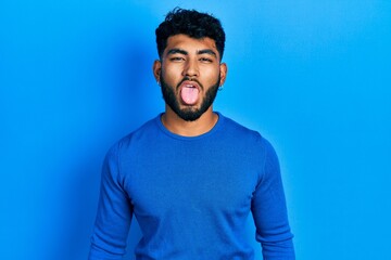 Fototapeta na wymiar Arab man with beard wearing casual blue sweater sticking tongue out happy with funny expression. emotion concept.