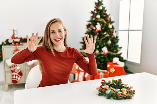 Caucasian young blonde woman sitting on the table by christmas tree showing and pointing up with fingers number nine while smiling confident and happy.