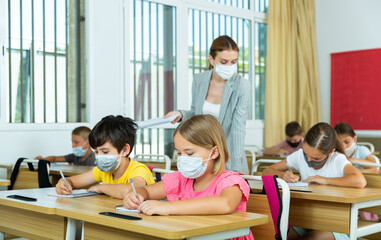 Fototapeta na wymiar Focused preteen pupils in protective face masks studying in classroom with female teacher. Necessary precautions in coronavirus pandemic