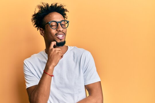Young african american man with beard wearing casual white t shirt and glasses thinking concentrated about doubt with finger on chin and looking up wondering