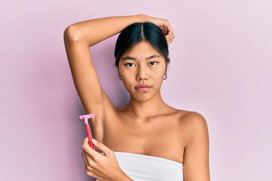 Young chinese woman wearing shower towel using razor relaxed with serious expression on face. simple and natural looking at the camera.