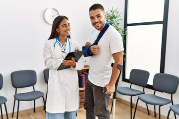 Fototapeta premium Youg latin injuried man wearing sling and using crutches talking with the doctor at clinic waiting room.