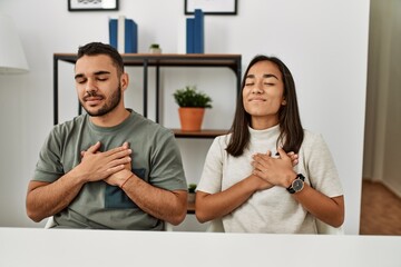 Young latin couple wearing casual clothes sitting on the table smiling with hands on chest with...