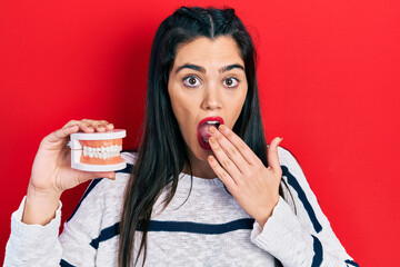 Young hispanic girl holding denture covering mouth with hand, shocked and afraid for mistake....