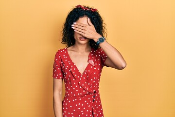 Young latin girl wearing summer dress covering eyes with hand, looking serious and sad. sightless, hiding and rejection concept