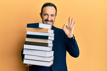 Middle age hispanic man holding a pile of books doing ok sign with fingers, smiling friendly gesturing excellent symbol