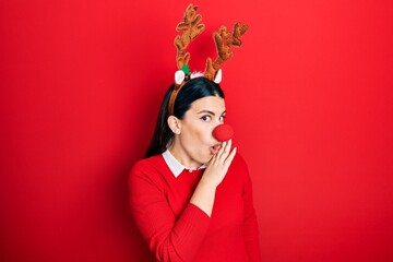 Young hispanic woman wearing deer christmas hat and red nose hand on mouth telling secret rumor, whispering malicious talk conversation
