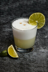 Glass of pisco sour. White drink in glass tumbler with ice and lemon on black marble background.
