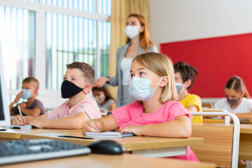 Fototapeta na wymiar Portrait of cute little schoolgirl in protective face mask studying in classroom. Necessary precautions in coronavirus pandemic..