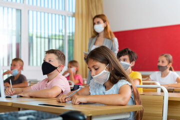 Fototapeta na wymiar Portrait of schoolgirl in protective face mask who writing exercises at lesson in primary school