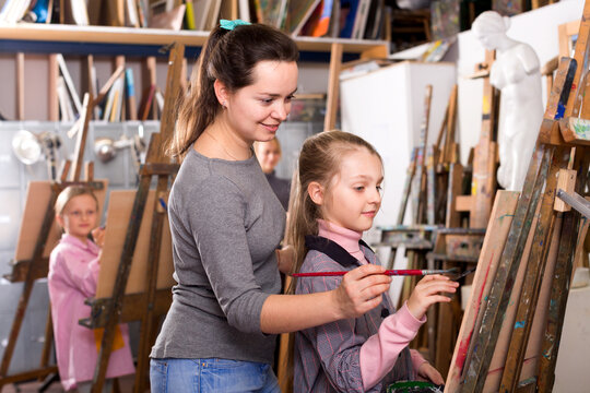 Beautiful young female teacher helping girl during painting class at art studio