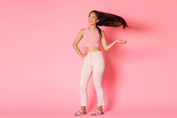 Obraz na płótnie Canvas Fashion, beauty and lifestyle concept. Stylish and carefree beautiful asian girl tossing her hair and looking confident aside, smiling pleased, enjoying vacation or shopping, standing pink background