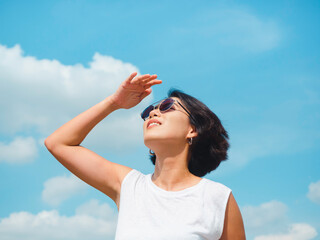 Woman in summertime. Smiling beautiful Asian woman short hair wearing sunglasses and white sleeveless shirt looking up and shading eyes with her hand on blue sky background on sunny day in summer. - Powered by Adobe