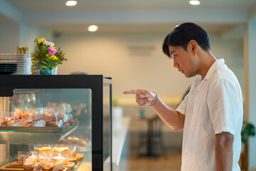 Asian man looking and choosing variation of delicious cake in bakery showcase fridge at cafe. Male customer buying tasty sweet dessert at coffee shop. Small business restaurant food and drink concept