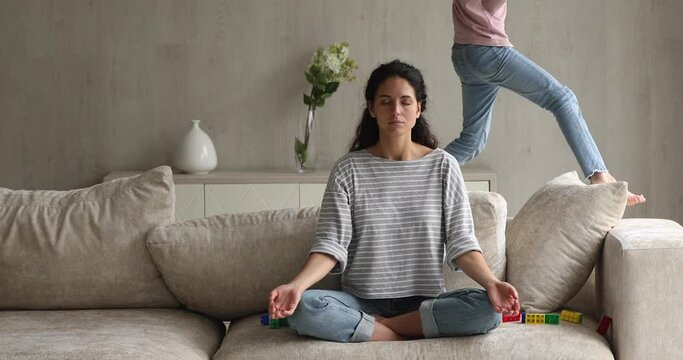 Peaceful young hispanic latin woman meditating on comfortable sofa in lotus position with closed eyes involved in yoga breathing exercises while carefree little child daughter playing running around.