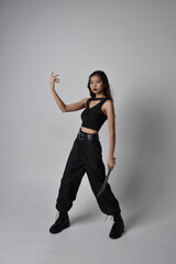 Full length portrait of pretty young asian girl wearing black tank top, utilitarian  pants and leather boots. Standing pose holding a knife,  isolated against a  studio background.
