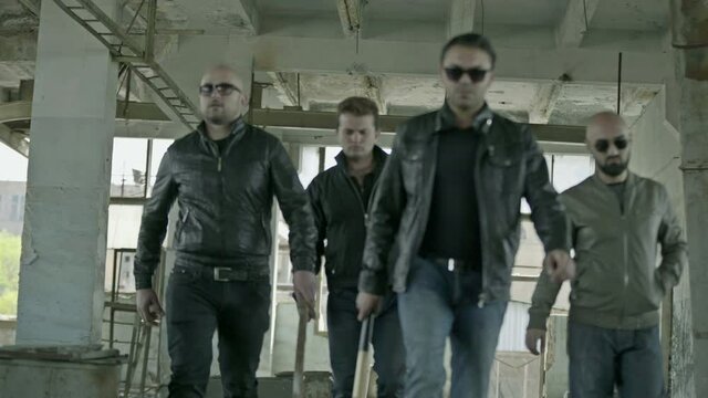 Defocused front view of four men in leather jackets confidently walking to camera in empty abandoned building . Organized criminal street gangsters with bat before fight . 