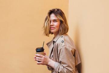 Young millennial woman with wild hair dressed in an autumn coat standing a cup of coffee to go near the wall.