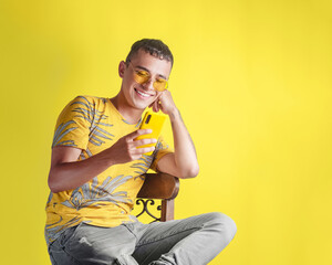 Handsome young man sitting on a chair while happily checking his cell phone on a yellow background