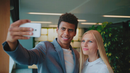 Cheerful black man with laughing blond woman taking selfie with smartphone in modern office having fun.