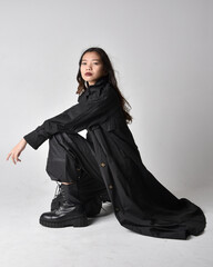 Naklejka premium Full length portrait of pretty young asian girl wearing black tank top, utilitarian pants, leather boots and large dark coat. Sitting pose with gestural hand movements, isolated on studio backgroun
