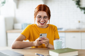 Smiling happy young woman with red hair texting on mobile phone sitting at home in the kitchen. Chatting with friends, relax at home