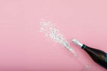 From above of bottle of champagne and glass goblet placed on table with silver glitter showing sparkles on pink background