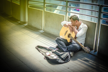 Fototapeta na wymiar Unemployed man in white shirt, sitting a long the overpass feel very tired. He is falling a sleep with his guitar. The global economic crisis from COVID-19 concept.
