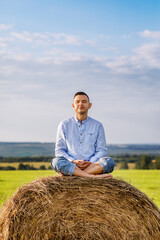 Naklejka na ściany i meble A middle-aged man sits in a yoga pose on a bale of hay. Summer vacation on the farm. Yoga classes in nature.