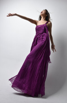 Full Length  Portrait Of Pretty Brunette Asian Girl Wearing Purple Flowing  Gown. Standing Pose In Low Angle On On Studio Background.