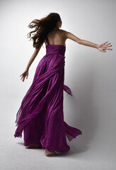 Full length  portrait of pretty brunette asian girl wearing purple flowing  gown. Standing pose in low angle on on studio background.