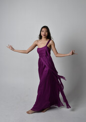 Full length  portrait of pretty brunette asian girl wearing purple flowing  gown. Standing, dancing pose on on studio background.